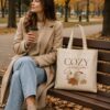 Woman sitting on a park bench with the Cozy Season tote and a coffee